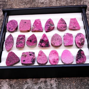 May include: A collection of polished, pink gemstones with dark inclusions, displayed in a black tray with white inserts. The stones vary in shape, including ovals, teardrops, and rectangles. The gemstones are arranged neatly, showcasing their unique textures and colors.