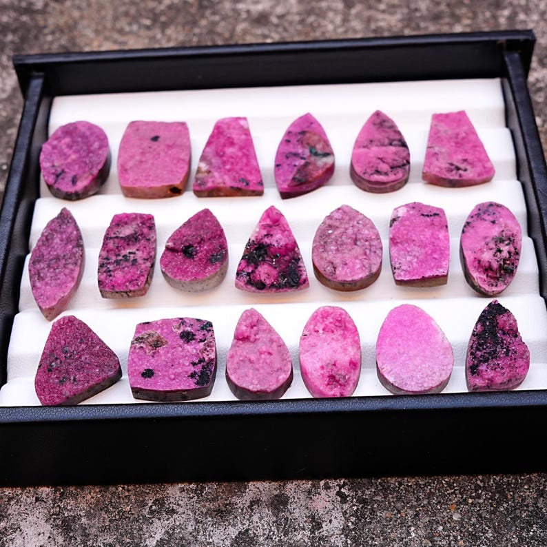 May include: A collection of polished, pink gemstones with dark inclusions, displayed in a black tray with white inserts. The stones vary in shape, including ovals, teardrops, and rectangles. The gemstones are arranged neatly, showcasing their unique textures and colors.