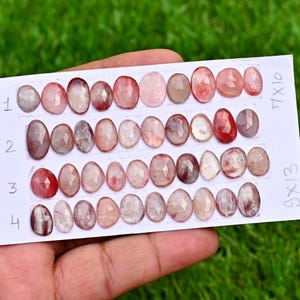 May include: A collection of oval-shaped gemstones in shades of pink, red, and clear. The gems are arranged on a white card with handwritten measurements of 7x10 and 9x13. The background is green grass.
