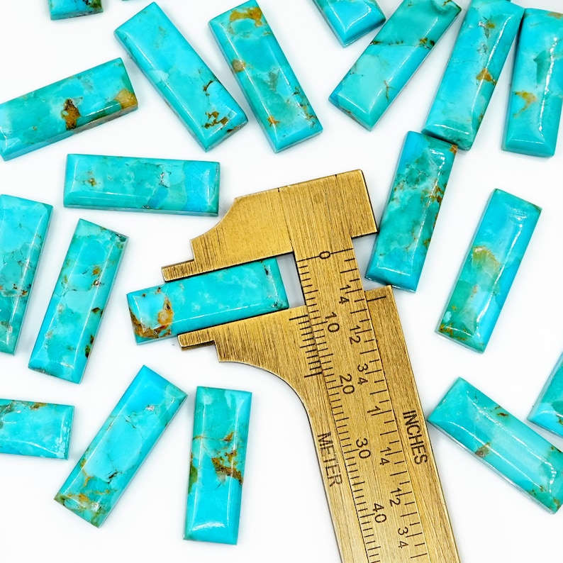May include: Rectangular turquoise gemstones with brown matrix patterns are scattered on a white surface. A brass ruler, marked in inches, is used to measure one of the stones. The gemstones are a vibrant blue-green color.