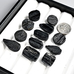 May include: A collection of raw, black tourmaline gemstones in various shapes and sizes, displayed in a black tray with white inserts. A US dime is included for size reference. The gemstones have a rough, textured surface.