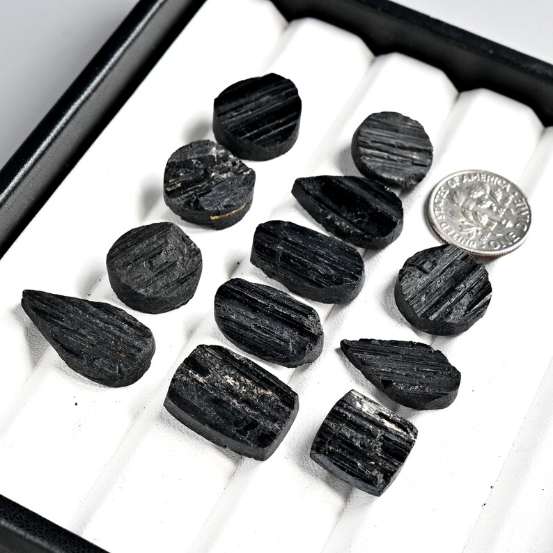 May include: A collection of raw, black tourmaline gemstones in various shapes and sizes, displayed in a black tray with white inserts. A US dime is included for size reference. The gemstones have a rough, textured surface.