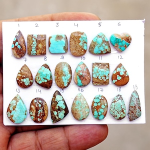 Puede incluir: Una colecci&oacute;n de diecinueve piedras preciosas de turquesa pulida dispuestas sobre una tarjeta blanca, numeradas del uno al diecinueve. Las piedras presentan formas variadas como l&aacute;grima, &oacute;valo, cuadrado y coraz&oacute;n, con patrones azul-verdosos y matriz marr&oacute;n.