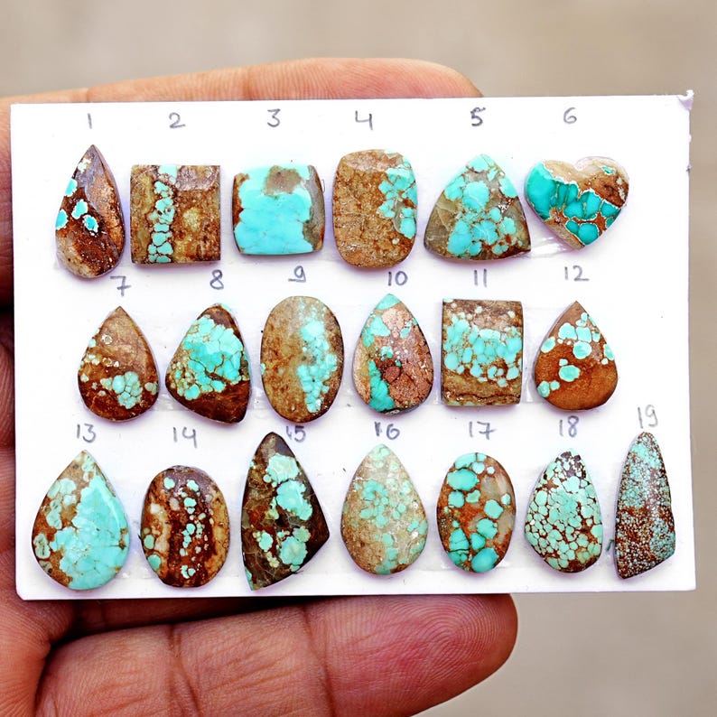 Puede incluir: Una colecci&oacute;n de diecinueve piedras preciosas de turquesa pulida dispuestas sobre una tarjeta blanca, numeradas del uno al diecinueve. Las piedras presentan formas variadas como l&aacute;grima, &oacute;valo, cuadrado y coraz&oacute;n, con patrones azul-verdosos y matriz marr&oacute;n.
