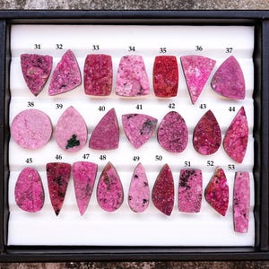 May include: A collection of polished, pink rhodonite gemstones in various shapes, including triangles, teardrops, and rectangles. The gemstones are displayed in a black tray with a white insert, each piece numbered for identification.
