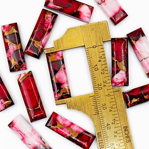 May include: Rectangular red and pink gemstone beads with gold veining are scattered around a brass ruler. The ruler shows measurements in inches. The beads have a polished, glossy finish, and vary in color from deep red to pale pink.