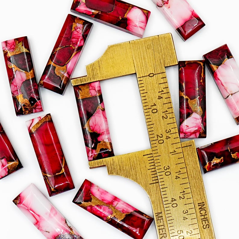 May include: Rectangular red and pink gemstone beads with gold veining are scattered around a brass ruler. The ruler shows measurements in inches. The beads have a polished, glossy finish, and vary in color from deep red to pale pink.