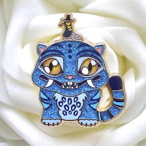 May include: A blue glitter enamel pin depicting a stylized tiger-like creature with large eyes, sharp teeth, and a small hat. The pin has gold-coloured metal accents and a detailed design, ideal for bags or jackets.