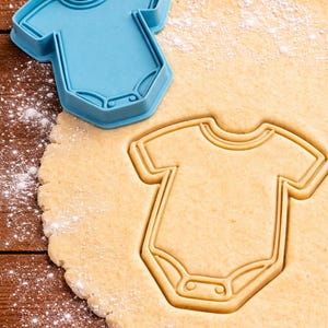 May include: A blue plastic onesie-shaped cookie cutter next to a cutout in dough. The dough is dusted with flour and sits on a wooden surface. The cookie cutter is a light blue colour and is next to the cutout of the same shape.