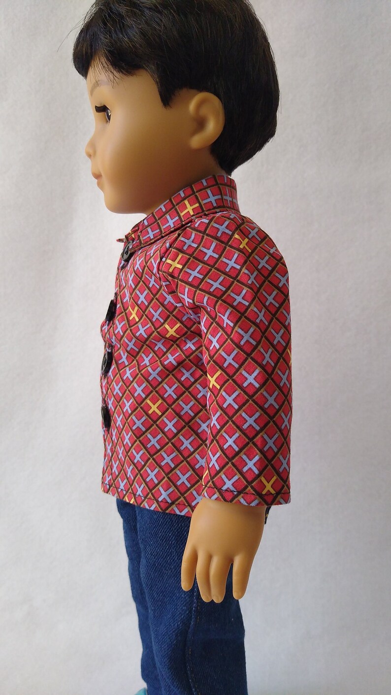 May include: A doll wearing a red, blue and yellow patterned shirt with a button-up closure and blue jeans.