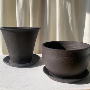 Pode incluir: Duas floreiras de cerâmica castanho-escuras com pires a condizer. Uma é um vaso alto e alargado, e a outra tem a forma de uma tigela arredondada. Ambas têm uma superfície texturizada e são adequadas para uso interior ou exterior.