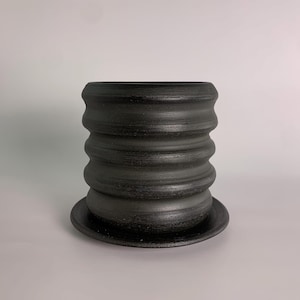 May include: A black ceramic planter with a textured, ridged design. The planter has a cylindrical shape with a matching saucer base. The dark color and unique texture give it a modern, minimalist aesthetic, suitable for indoor or outdoor use.
