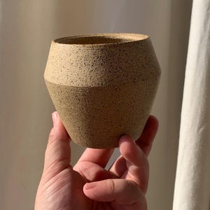 May include: A handcrafted, speckled ceramic vase with a two-tone design. The upper portion is a lighter tan color, while the lower half is a darker tan. The vase has a wide opening and a geometric shape, perfect for displaying small plants or flowers.