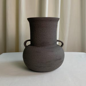 May include: A dark brown ceramic vase with a textured finish. The vase features a rounded base, a cylindrical neck, and two small handles. The vase is placed on a white surface.