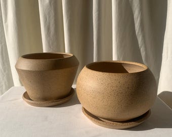 May include: Two ceramic plant pots with matching saucers. The pots are a speckled brown color and have a rounded shape. The smaller pot has a more angular shape.