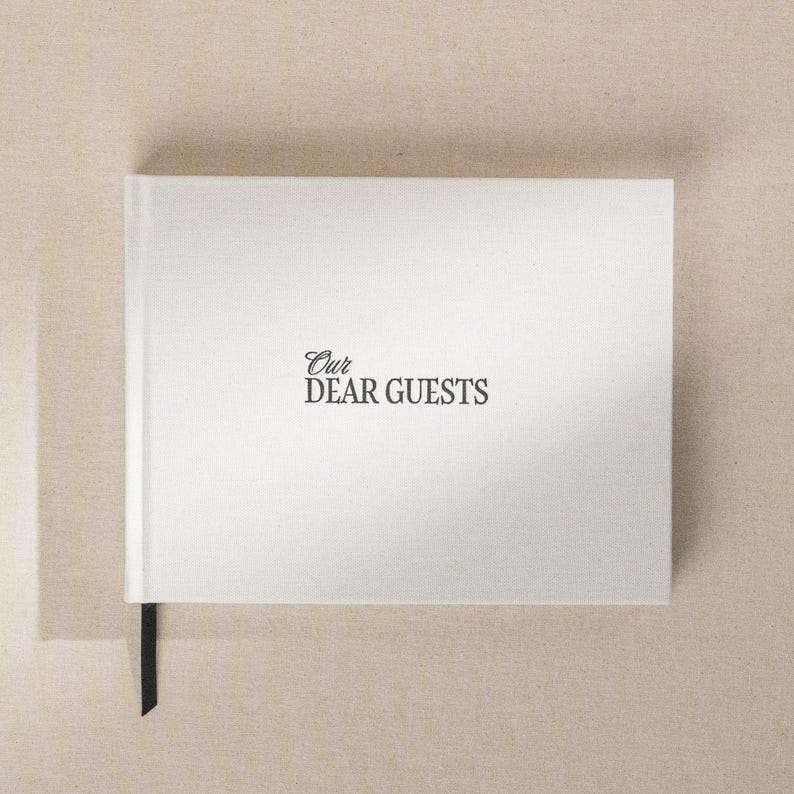May include: A white guest book with the text "Our Dear Guests" in a script font. The book has a black ribbon bookmark. The background is a neutral beige color.