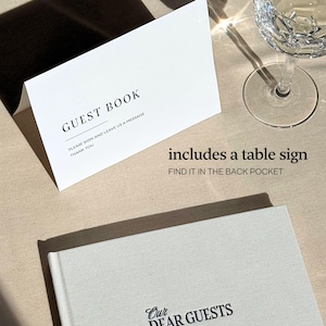 May include: A guest book and table sign set. The guest book is white with the words "Our Dear Guests" printed on the cover. A white table sign reads "GUEST BOOK" with instructions to sign and leave a message. A crystal wine glass is in the background.