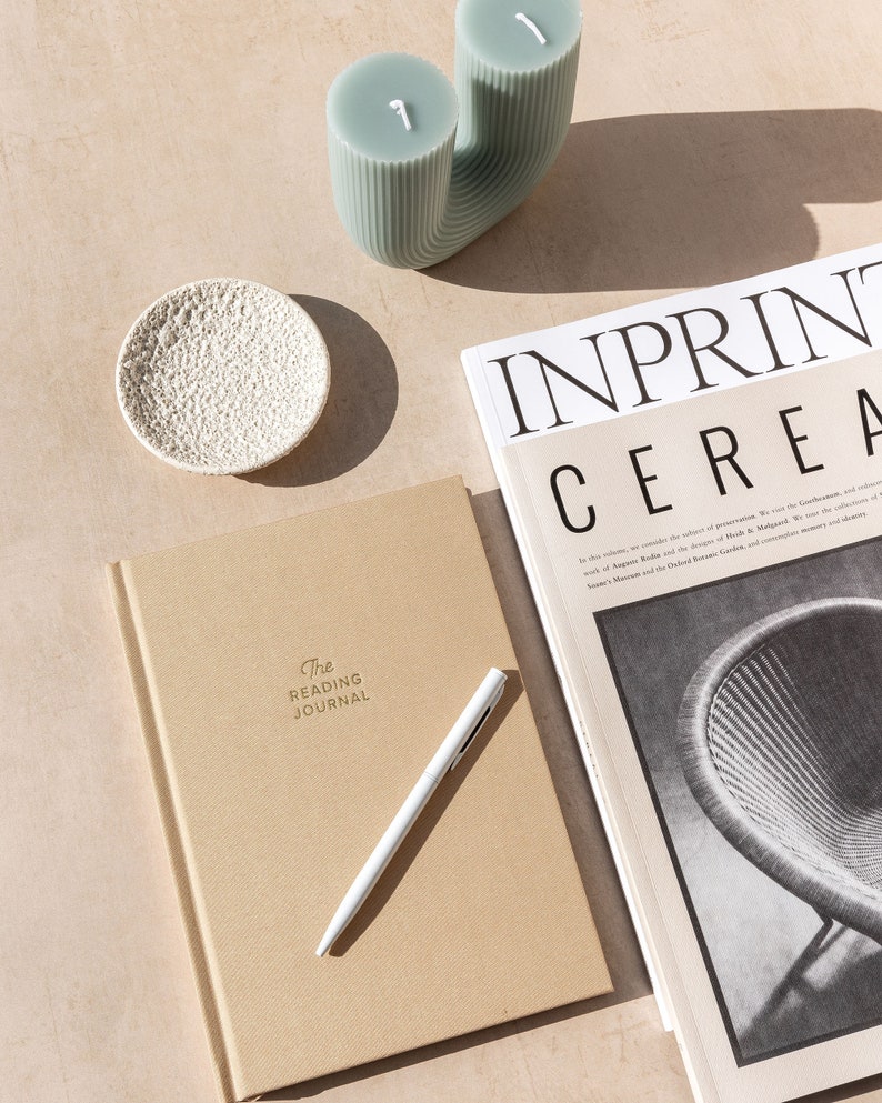 May include: A tan journal with the text "The Reading Journal" embossed on the cover. A white pen is lying on top of the journal. A magazine with the text "INPRINT CEREA" is visible in the background.
