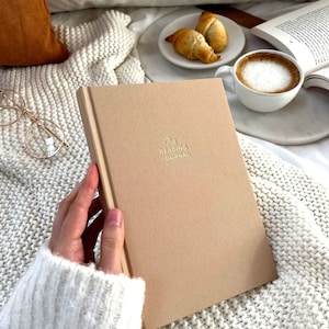 May include: A beige journal with the text "The Reading Journal" in gold lettering. The journal is on a white knitted blanket with a cup of coffee and croissants in the background.