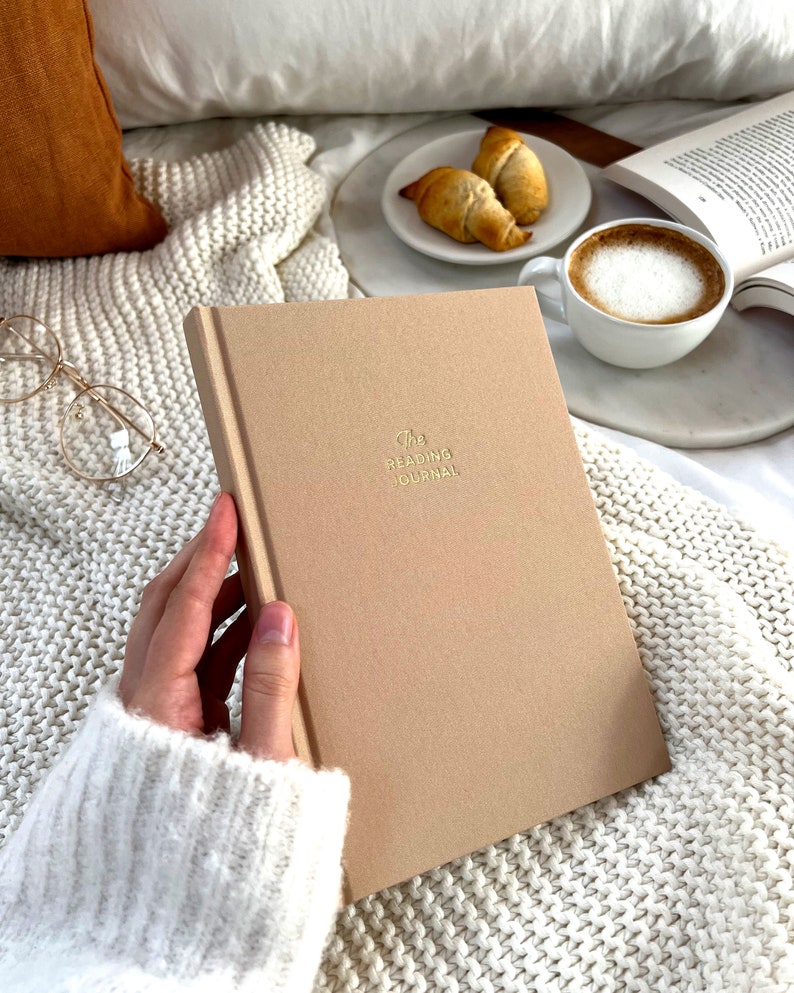 May include: A beige journal with the text "The Reading Journal" in gold lettering. The journal is on a white knitted blanket with a cup of coffee and croissants in the background.