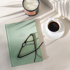 May include: A green journal with the text "The Reading Journal" on the cover. A pair of tortoiseshell glasses are lying on top of the journal. A white coffee cup with black coffee is on a white saucer. A white candle is in the background.