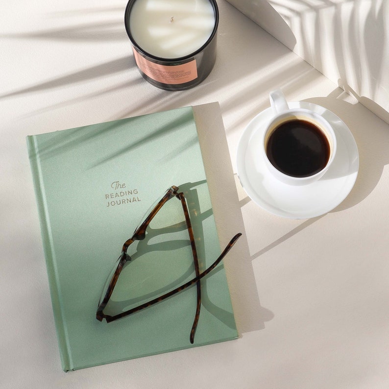 May include: A green journal with the text "The Reading Journal" on the cover. A pair of tortoiseshell glasses are lying on top of the journal. A white coffee cup with black coffee is on a white saucer. A white candle is in the background.