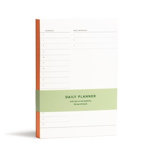 Puede incluir: Un bloc de notas de planificaci&oacute;n diaria con una cubierta verde y naranja. El bloc de notas tiene 100 hojas de papel que se pueden arrancar y dice "DAILY PLANNER" y "every day is a new beginning" en la cubierta.