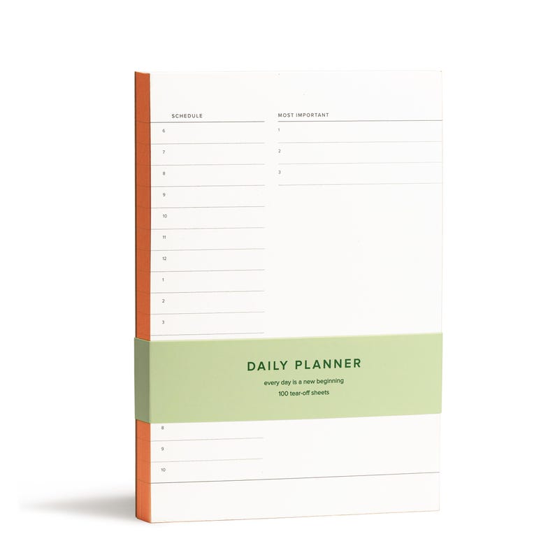 Puede incluir: Un bloc de notas de planificaci&oacute;n diaria con una cubierta verde y naranja. El bloc de notas tiene 100 hojas de papel que se pueden arrancar y dice "DAILY PLANNER" y "every day is a new beginning" en la cubierta.