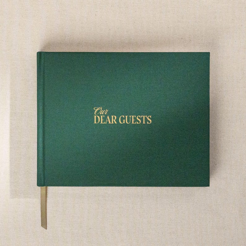 May include: A dark green guest book with the gold text "Our Dear Guests" on the cover. The book has a matching green ribbon bookmark. The book is rectangular and has a textured cover.