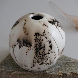 May include: A round, cream-coloured ceramic vase with a single opening. The vase features black, lightning-like patterns and dark shading. The vase rests on a grey stone surface. A dried plant is in the background.