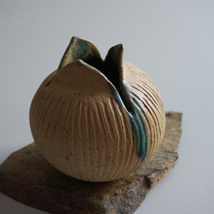 May include: A round ceramic vase with a textured surface and a teal glaze. The vase has a unique opening at the top, resembling a flower.