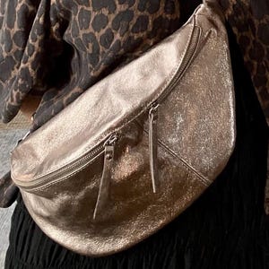 May include: A bronze-coloured fanny pack with a curved design and two zipped compartments. The bag features a matching strap and a silver-tone clasp. The background includes a leopard print top and a black skirt.
