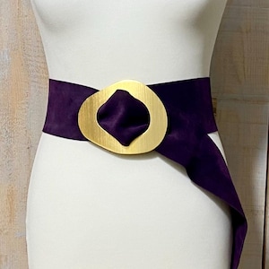 May include: A mannequin displays a wide, deep purple belt with a large, gold-colored buckle. A black and white spotted scarf is draped around the mannequin's neck. The belt is the focal point, showcasing a bold fashion statement.