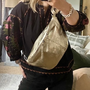 Könnte beinhalten: Eine goldfarbene Metallic-Bauchtasche mit Reißverschluss, quer über dem Körper getragen. Die Tasche wird mit einem schwarzen Oberteil mit bestickten Ärmeln und einer schwarzen Hose kombiniert. Das Bild wird in einem Spiegel aufgenommen.