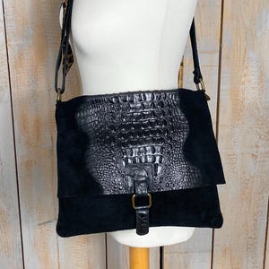 Könnte beinhalten: Schwarze Leder-Umhängetasche mit Klappenverschluss und strukturiertem Krokodilmuster. Die Tasche hat einen Wildleder-Körper und eine goldfarbene Schnalle. Ein Leoparden-Schal ist über der Schaufensterpuppe drapiert.