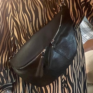 May include: Black leather fanny pack with a silver zipper and tassel detail. The bag is worn over a long-sleeved dress with a zebra stripe pattern in black and tan.