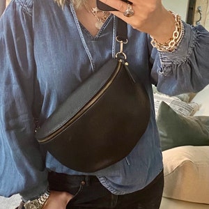 May include: A black leather fanny pack with a gold zipper is worn across the body. The person is wearing a denim blue shirt and black jeans. A gold chain bracelet and watch are visible.