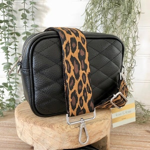 Op de afbeelding: Zwarte leren crossbodytas met een gewatteerd patroon en een afneembare luipaardprint riem. De tas heeft zilverkleurige hardware en een ritssluiting. De tas wordt gepresenteerd op een houten standaard.