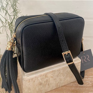 Puede incluir: Bolso bandolera de cuero negro con cremallera y hebilla doradas. El bolso tiene una superficie texturizada y una correa larga ajustable. Una borla negra cuelga del lateral, con una etiqueta "RR".
