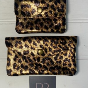 May include: Two small, rectangular leopard print pouches with a gold metallic finish. Each pouch has a flap closure secured by a single, round, brass-coloured button. The pouches are set against a white wooden background.