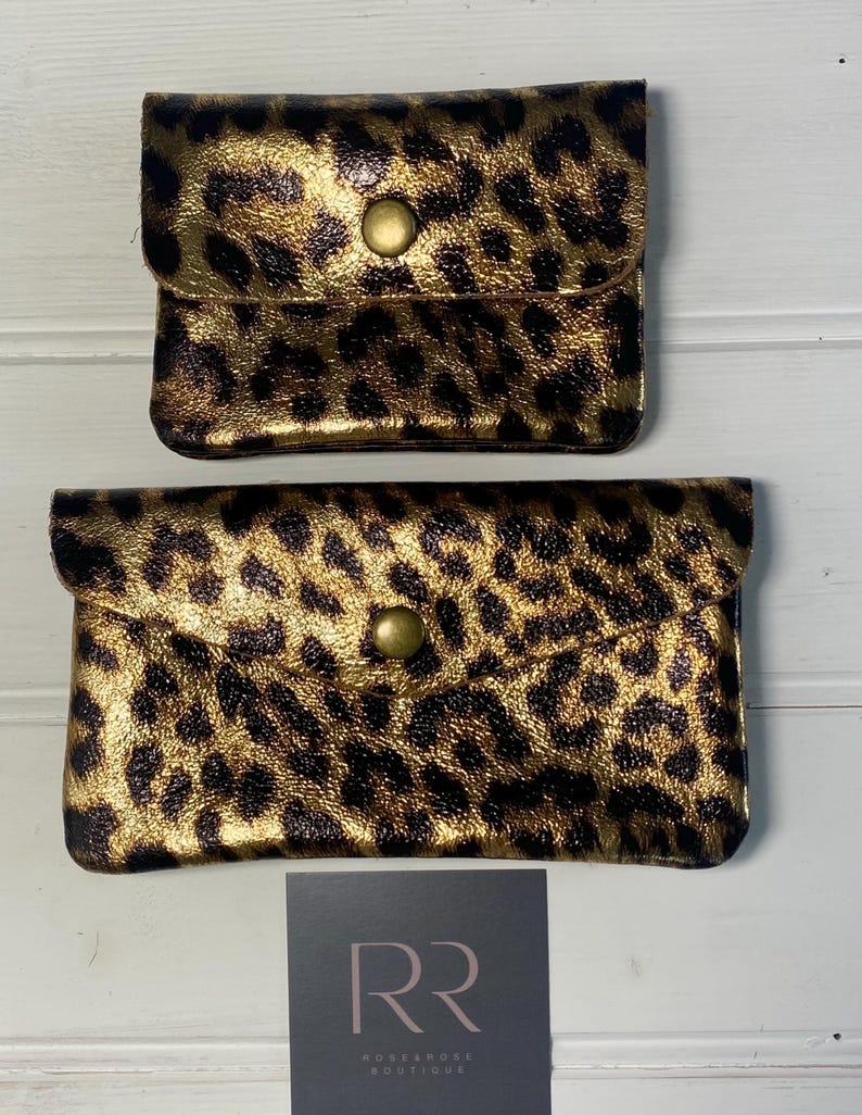 May include: Two small, rectangular leopard print pouches with a gold metallic finish. Each pouch has a flap closure secured by a single, round, brass-coloured button. The pouches are set against a white wooden background.