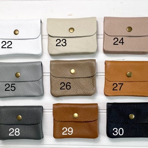 May include: A collection of small, square leather wallets in various colors, including white, beige, taupe, gray, brown, and black. Each wallet has a flap closure secured by a gold-colored snap. Numbers 22 through 30 are printed on the wallets.