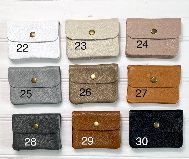 May include: A collection of small, square leather wallets in various colors, including white, beige, taupe, gray, brown, and black. Each wallet has a flap closure secured by a gold-colored snap. Numbers 22 through 30 are printed on the wallets.