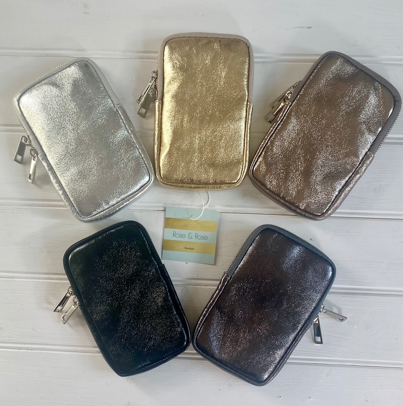 May include: Five metallic zippered phone cases in silver, gold, bronze, black, and dark brown are arranged on a white wooden surface. Each case has a rectangular shape with a zipper closure and a small tag that reads "Rose & Rose Boutique."