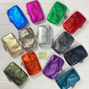 May include: A collection of metallic-finish phone cases in various colors, including blue, pink, green, red, gold, silver, black, orange, purple, and teal. Each case has a zipper closure and a rectangular shape. The cases are arranged on a white wooden surface.