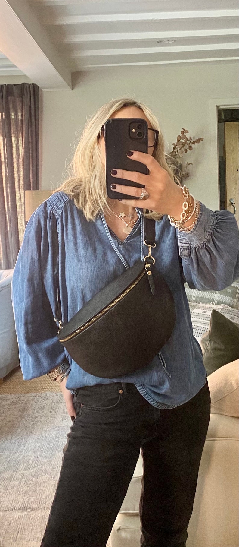 May include: Black leather fanny pack with a gold zipper worn across the body. The bag is paired with a blue denim shirt and black jeans. The person is wearing gold bracelets and a necklace.