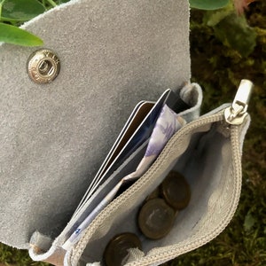 May include: A light grey suede leather wallet, open to show cards, cash, and coins. The wallet features a snap closure and a zipped coin pouch. The interior lining is a lighter shade of grey. Designed for carrying essential items.