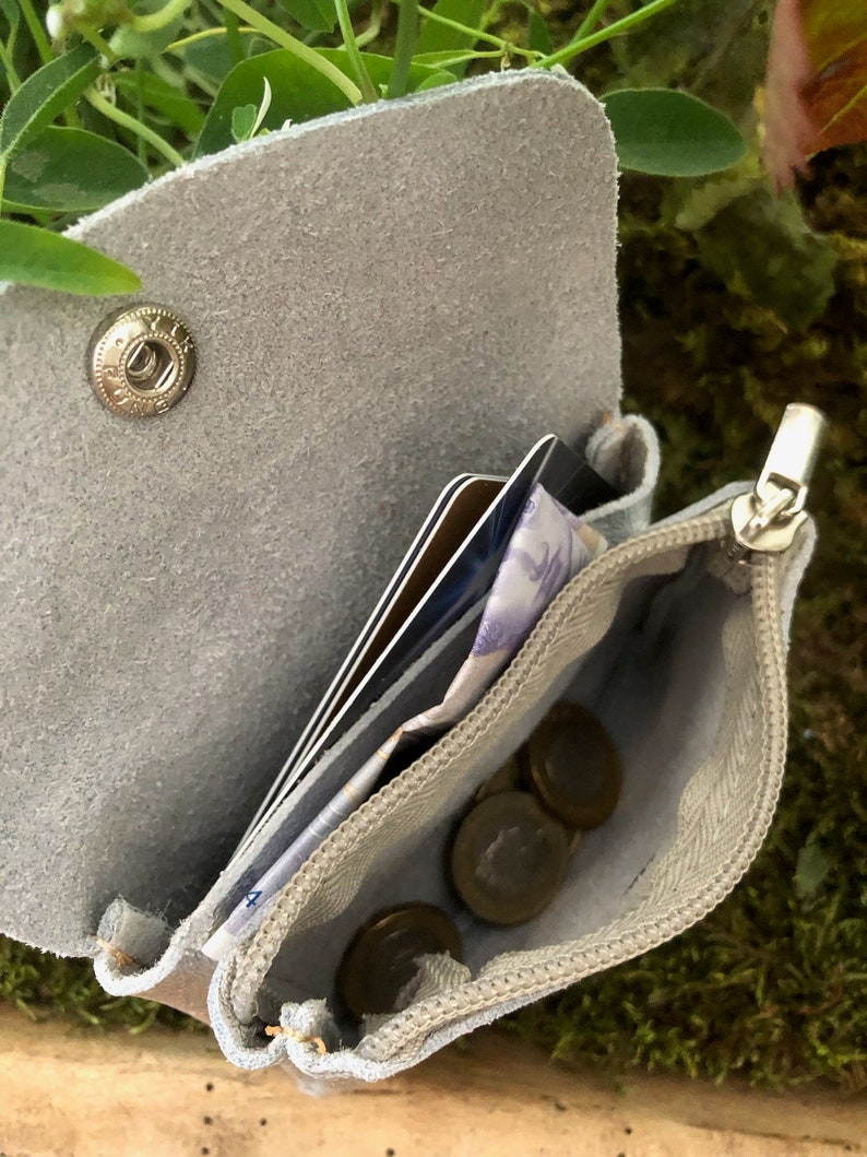 May include: A light grey suede leather wallet, open to show cards, cash, and coins. The wallet features a snap closure and a zipped coin pouch. The interior lining is a lighter shade of grey. Designed for carrying essential items.