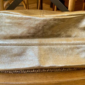 May include: A gold-colored leather tote bag with a rectangular shape. The bag has a stitched seam detail across the middle and a black strap. The bag is sitting on a woven surface.