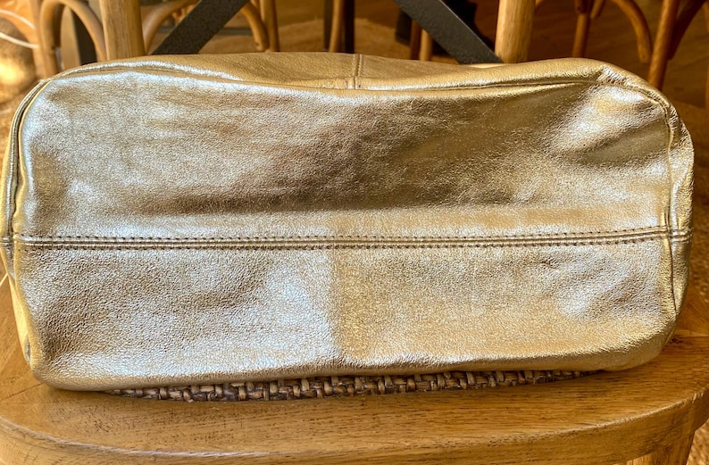 May include: A gold-colored leather tote bag with a rectangular shape. The bag has a stitched seam detail across the middle and a black strap. The bag is sitting on a woven surface.